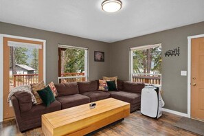 Un salon avec un canapé d’angle marron, une table basse en bois et un radiateur blanc. De grandes fenêtres offrent une vue sur les arbres et donnent sur une terrasse.