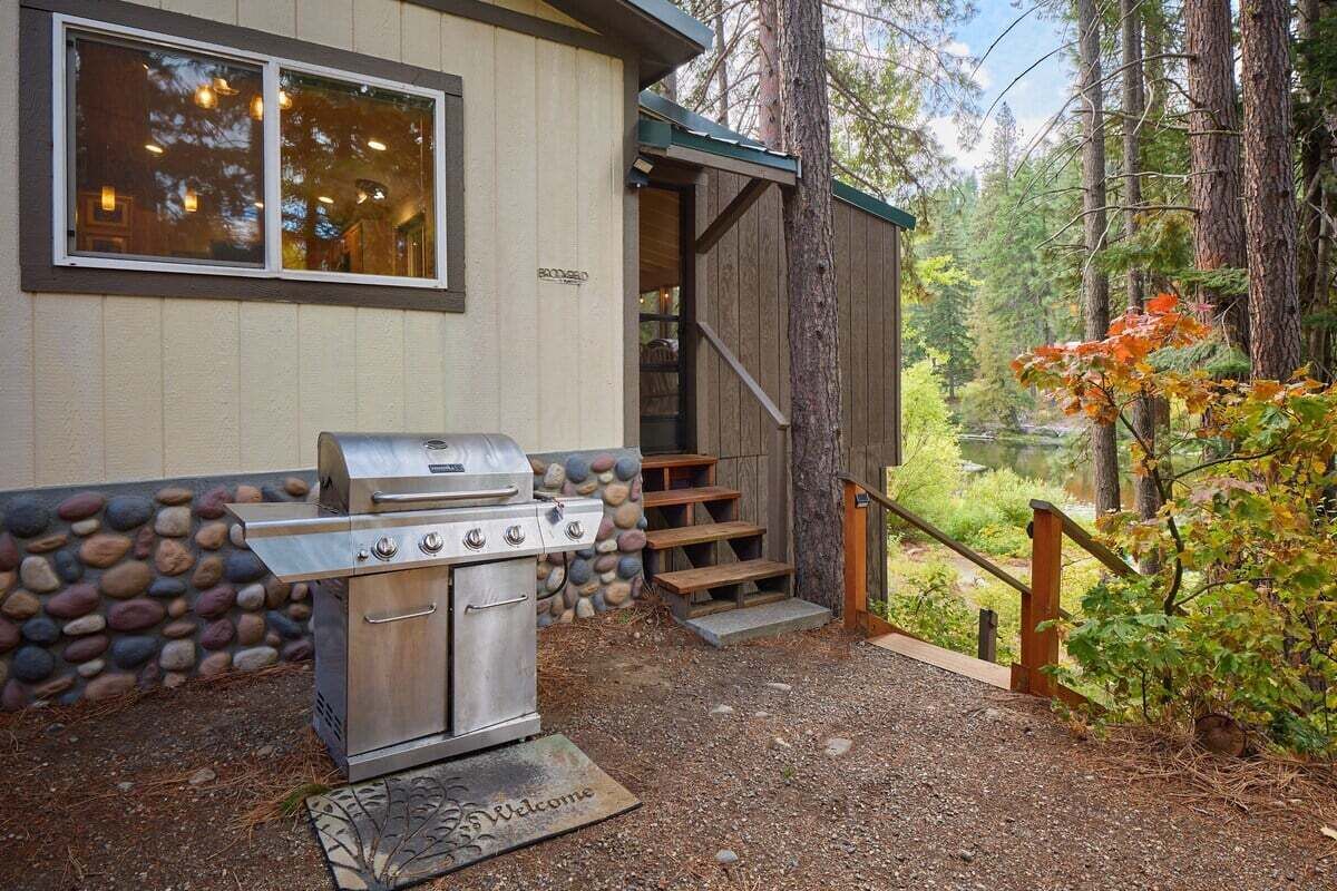Una parrilla de barbacoa frente a un edificio con pared de piedra, escalones de madera y vista a un bosque.
