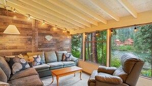 Un salón acogedor con sofá, sillón y mesa de centro, ventanas amplias con vistas a un bosque y una cabaña, y vigas de madera en el techo.