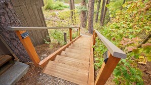 Una escalera de madera que conduce a una terraza de madera con barandilla de metal, rodeada de árboles y un paisaje natural.