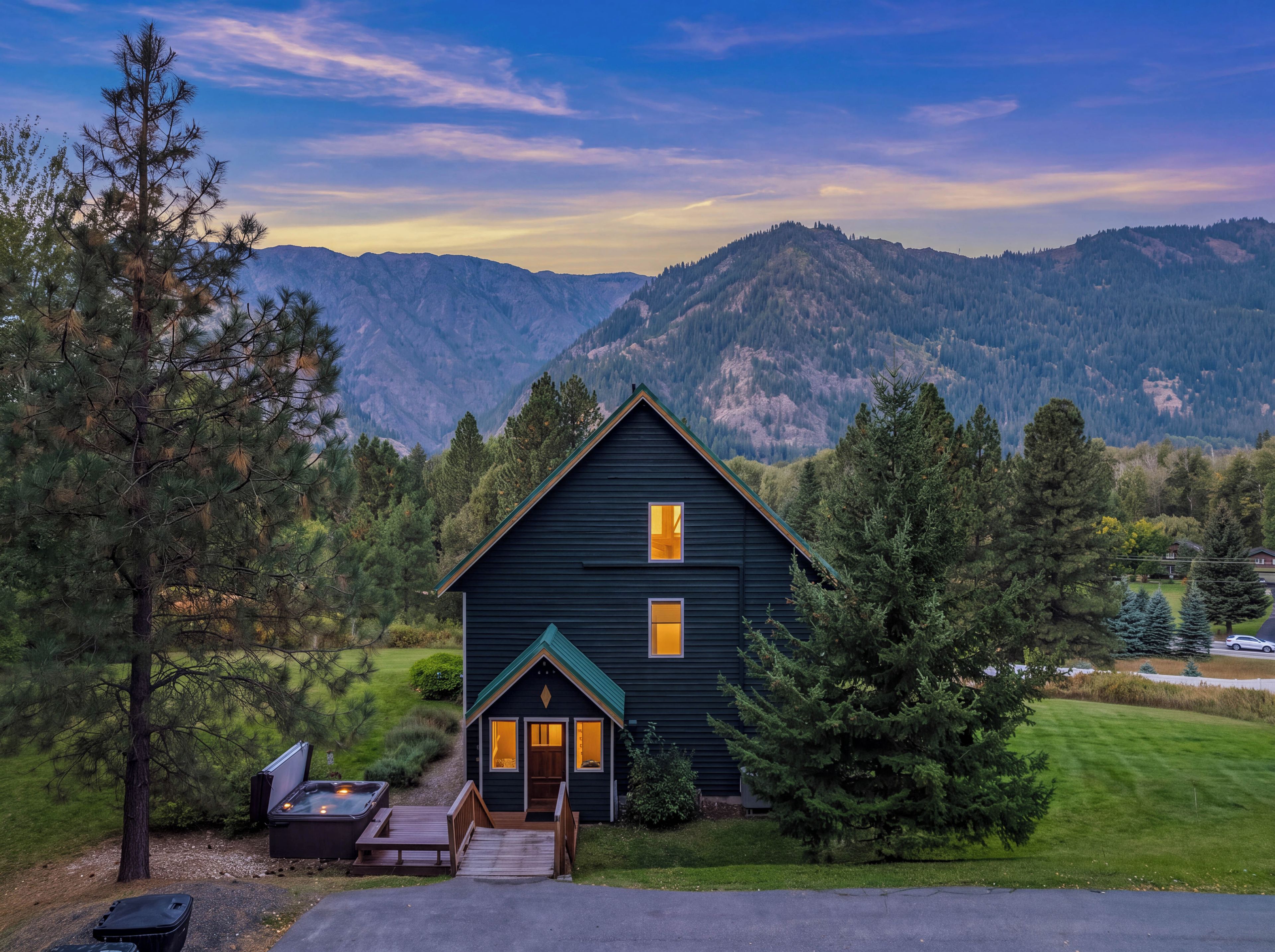 A dark-colored cabin with a deck and hot tub, surrounded by trees and mountains.