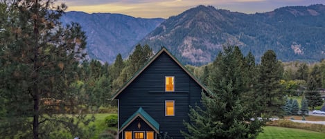 A dark-colored cabin with a deck and hot tub, surrounded by trees and mountains.