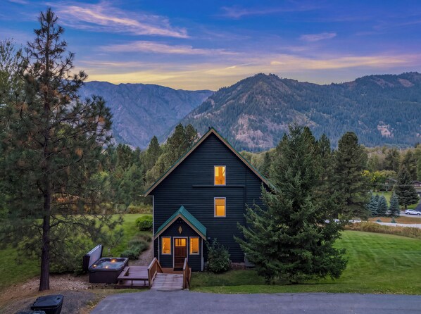 A dark-colored cabin with a deck and hot tub, surrounded by trees and mountains.