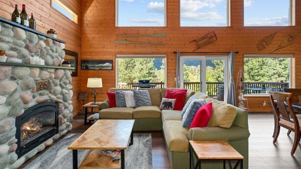 Un intérieur de chalet chaleureux avec une cheminée en pierre, des murs en bois, un canapé d’angle et de grandes fenêtres offrant une vue sur l’extérieur.