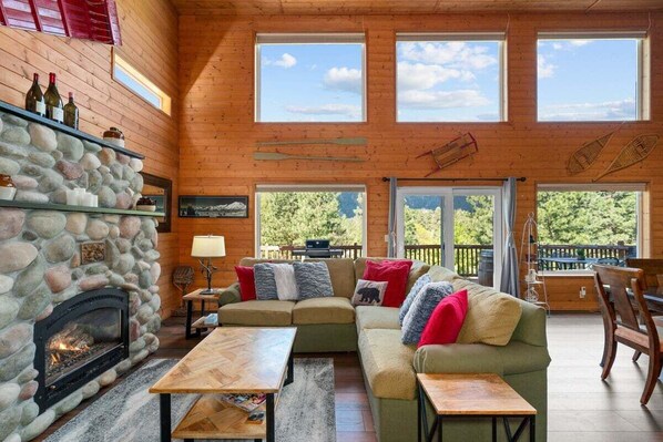 A cozy cabin interior with a stone fireplace, wooden walls, a sectional sofa, and large windows offering views of the outdoors.