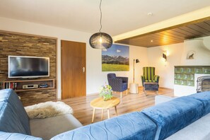 Ein modernes Wohnzimmer mit einem Flachbildfernseher, einem großen blauen Sofa, einem gemusterten Sessel und einer Holzbalkendecke.