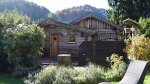 A rustic log cabin with a wooden door and window, surrounded by a garden with a bench and a chair.