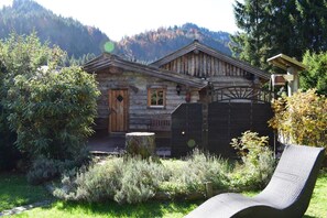 Ein rustikales Blockhaus mit Holztür und -fenster, umgeben von einem Garten mit Bank und Stuhl.