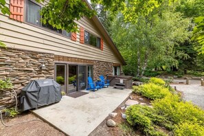 Una casa de dos pisos con fachada de piedra, una parrilla cubierta, mobiliario exterior y un jardín.