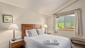 Habitación de hotel con cama, dos lámparas de noche, ventana con cortinas, cabecero de madera y un cuadro colgado en la pared.