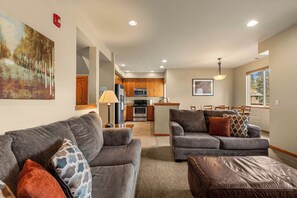 A modern living room with grey sofas and a brown ottoman, a kitchen with wooden cabinets and stainless steel appliances, and a dining area with a table and chairs.