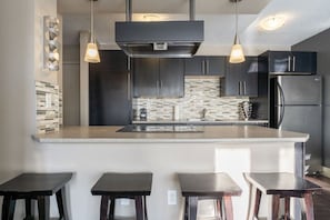 A modern kitchen with a central island, dark cabinetry, a stainless steel refrigerator, and pendant lighting.