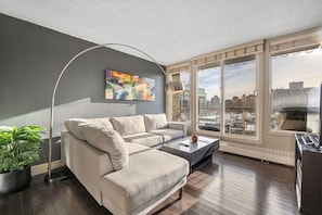 A modern living room with a sofa, coffee table, floor lamp, and a city view through large windows.