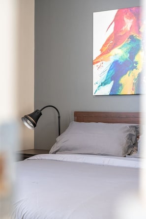 A bed with white bedding, a wooden headboard, a black floor lamp, and a colorful abstract painting on the wall.