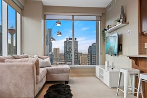 A modern living room with a large window, a sofa, a small table, and a TV mounted on the wall.