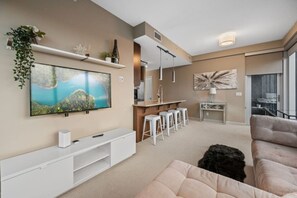 A modern living room with a TV, white entertainment unit, bar area with stools, and a large abstract painting.