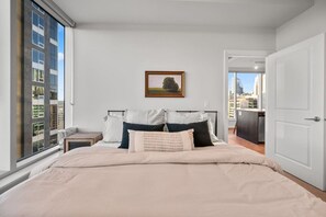 Quarto com uma cama grande, uma mesa de cabeceira, um quadro na parede e vista para a cidade através de uma janela.