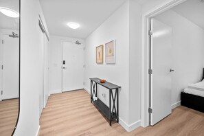 Een gang met een houten vloer, witte wanden en een zwarte console tafel met een klein oranje voorwerp erop. Er hangen afbeeldingen in lijsten aan de muur en bevindt zich een deur aan de rechterkant.