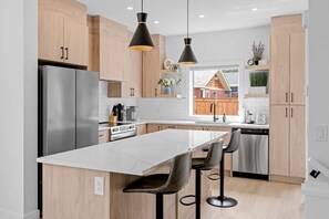 Una cocina moderna con armarios de madera, un refrigerador de acero inoxidable, una isla central con taburetes de bar, iluminación colgante y vistas a una terraza de madera a través de una ventana.