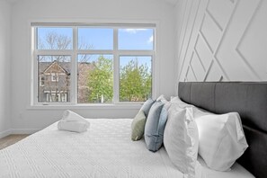 Un dormitorio con un ventanal grande, una cama con ropa blanca y un cabecero gris.