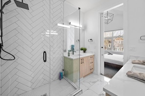 Un baño moderno con cabina de ducha de vidrio, tocador blanco con espejo y vista al dormitorio a través de una puerta abierta.