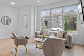 Una sala moderna con un sofá, sillas, una mesa de centro y un ventanal con vistas a casas.