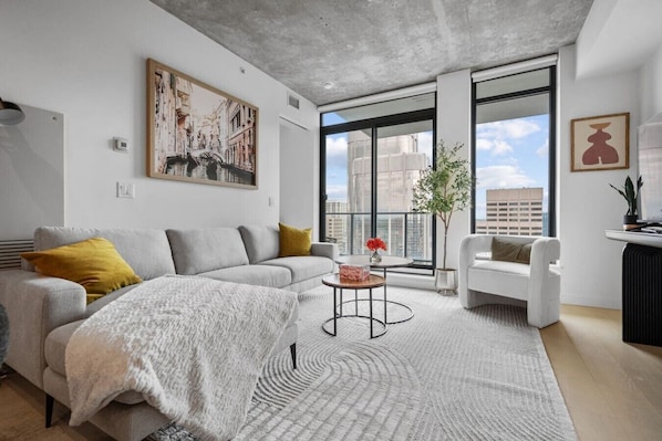 A modern living room with a grey sofa, a white armchair, a round coffee table, and a large window with a city view.