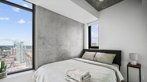 A modern bedroom with a large window, a bed with pillows, a bedside table with a lamp, and a view of the cityscape. - DT Views |UG Park | Mins to Saddledome | Gym (Calgary)