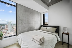 A modern bedroom with a large window, a bed with pillows, a bedside table with a lamp, and a view of the cityscape.
