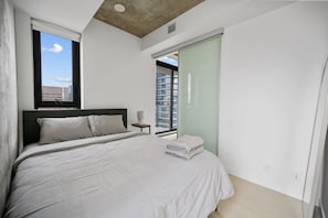 A bedroom with a bed, a nightstand, a window, and a view of buildings.