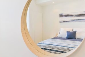 Quarto com cama, almofadas, uma obra de arte na parede e um espelho redondo.