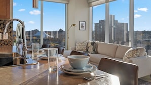 Une cuisine moderne dotée d’une grande fenêtre, un coin repas avec une table préparée pour deux personnes, et un canapé offrant une vue sur la ville.