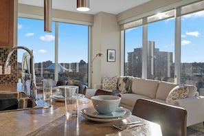Uma cozinha moderna com uma janela grande, uma área de refeições com mesa posta para dois e um sofá com vista para a cidade.