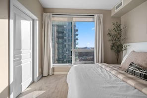 Quarto com uma janela ampla, cama com cabeceira, planta em vaso e vista para um edifício.