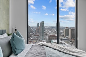 Quarto com uma janela grande, uma cama, um abajur e vista para a paisagem urbana.