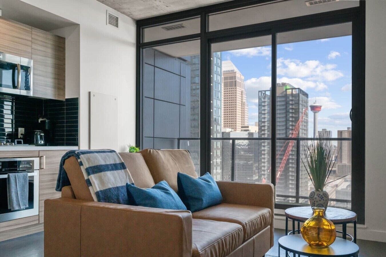 A modern living room with a sofa, a small table with a vase, and a view of a cityscape through large windows.