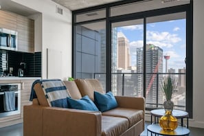 A modern living room with a sofa, a small table with a vase, and a view of a cityscape through large windows.