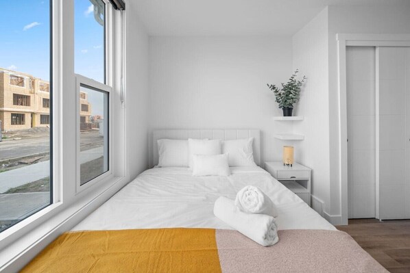 Une chambre d’hôtel moderne avec une grande fenêtre, un lit avec du linge de maison blanc, une table de chevet avec une lampe et une petite plante.