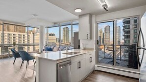 Une cuisine moderne dotée d’un grand îlot central, d’un coin repas avec des chaises et de grandes fenêtres offrant une vue sur la ville.