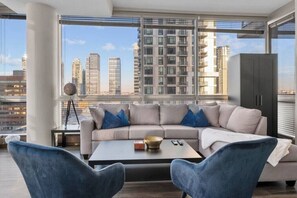 Een moderne woonkamer met een bank, twee fauteuils, een salontafel en een uitzicht op hoge gebouwen door grote ramen.