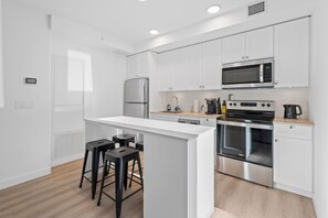 Una cocina moderna con armarios blancos, un refrigerador de acero inoxidable, un microondas, un horno y una cafetera. Hay tres taburetes negros en la isla de la cocina.