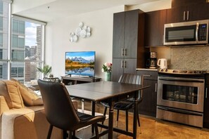 Cozinha moderna com armários escuros, micro-ondas de aço inoxidável e forno. Há uma mesa de jantar com cadeiras, uma TV de tela plana na parede e vista para a cidade através de grandes janelas.