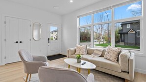 A modern living room with a sofa, two armchairs, a coffee table, and a large window offering a view of the neighborhood. - King Bed | Ping Pong | 3BR | Garage | AC |Backyard (Calgary)