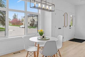 Une pièce avec une table ronde, des chaises blanches, un luminaire suspendu et une vue sur une rue et des maisons à l’extérieur.