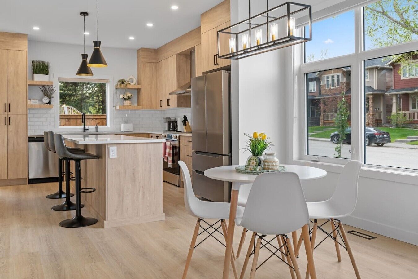 A modern kitchen with wooden cabinets, a central island, bar stools, a dining table with chairs, and a large window with a view of a residential street.