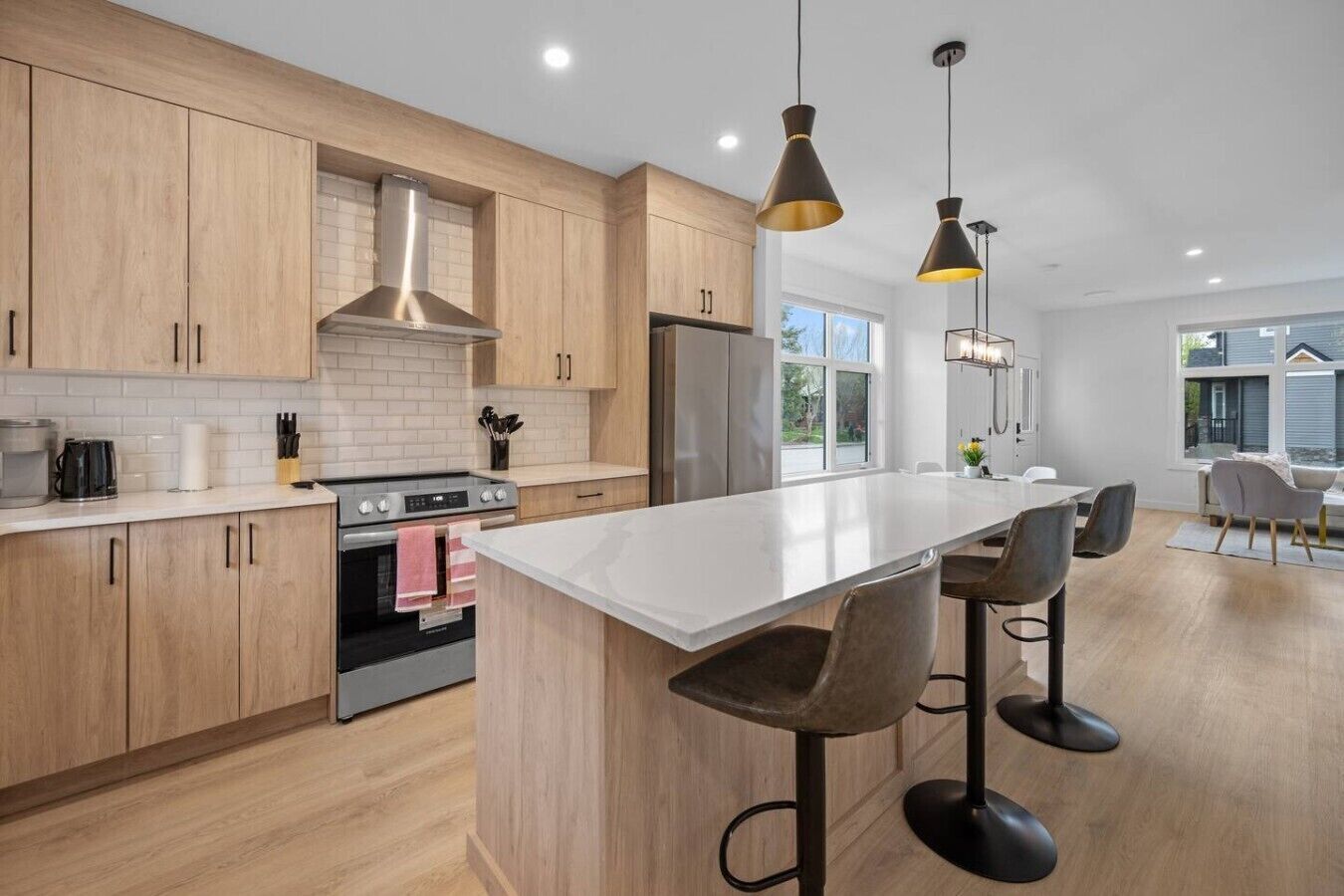 Une cuisine moderne avec des meubles en bois, un îlot central doté de tabourets de bar, une hotte de cuisine en acier inoxydable et des luminaires suspendus.