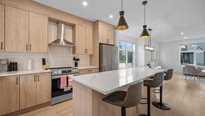 A modern kitchen with wooden cabinets, a central island with bar stools, a stainless steel range hood, and pendant lighting. - King Bed | Ping Pong | 3BR | Garage | AC |Backyard (Calgary)