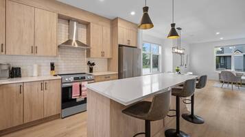 Une cuisine moderne avec des meubles en bois, un îlot central doté de tabourets de bar, une hotte de cuisine en acier inoxydable et des luminaires suspendus.