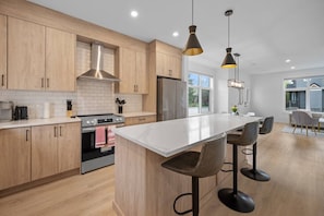 Une cuisine moderne avec des meubles en bois, un îlot central doté de tabourets de bar, une hotte de cuisine en acier inoxydable et des luminaires suspendus.