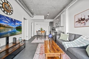 Una sala de estar moderna con un televisor de pantalla plana, una mesa de centro de madera, un sofá gris y una cocina al fondo.